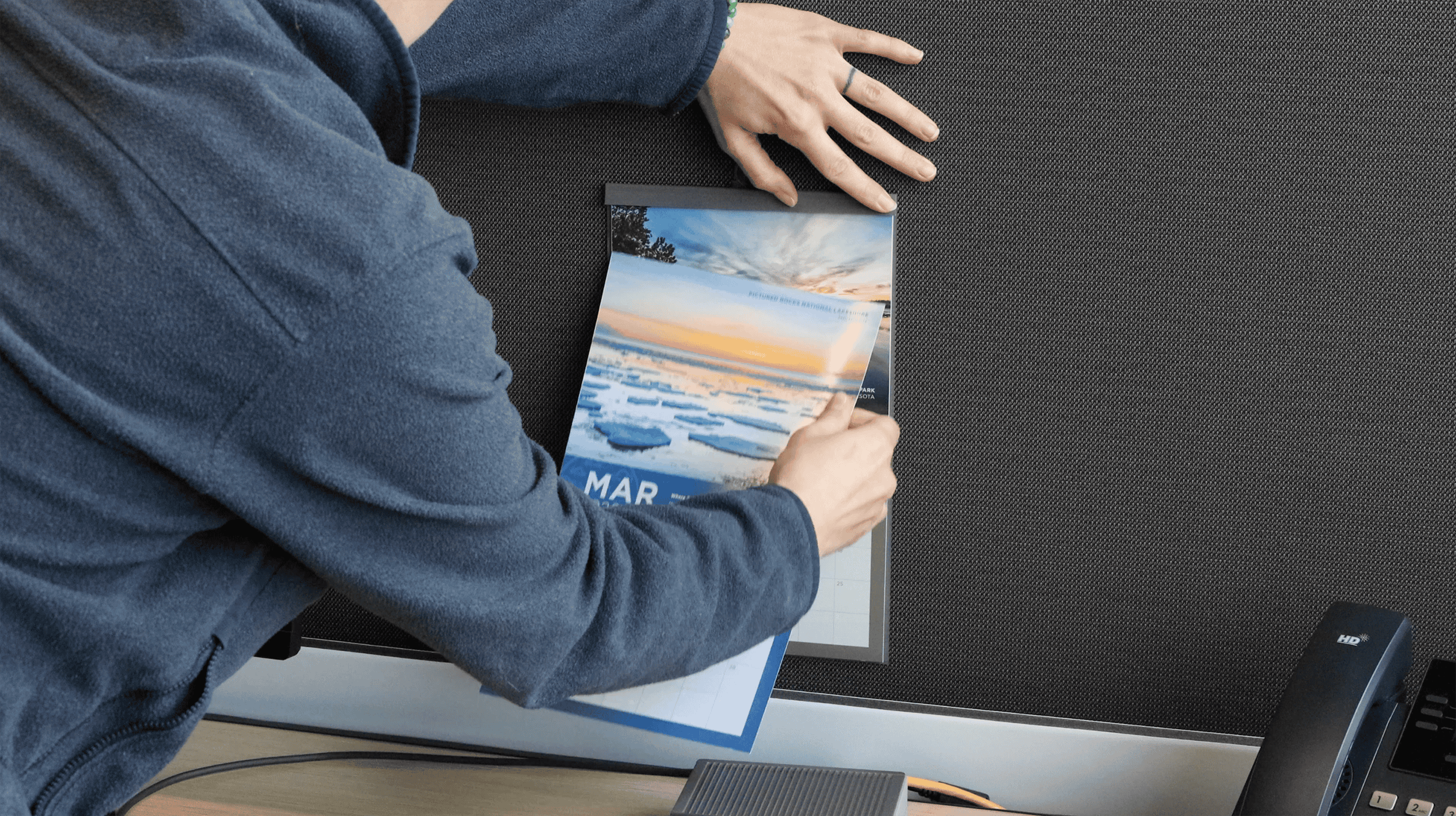 Person flipping the calendar to the March 2026 page, showing a winter shoreline at Pictured Rocks National Lakeshore.