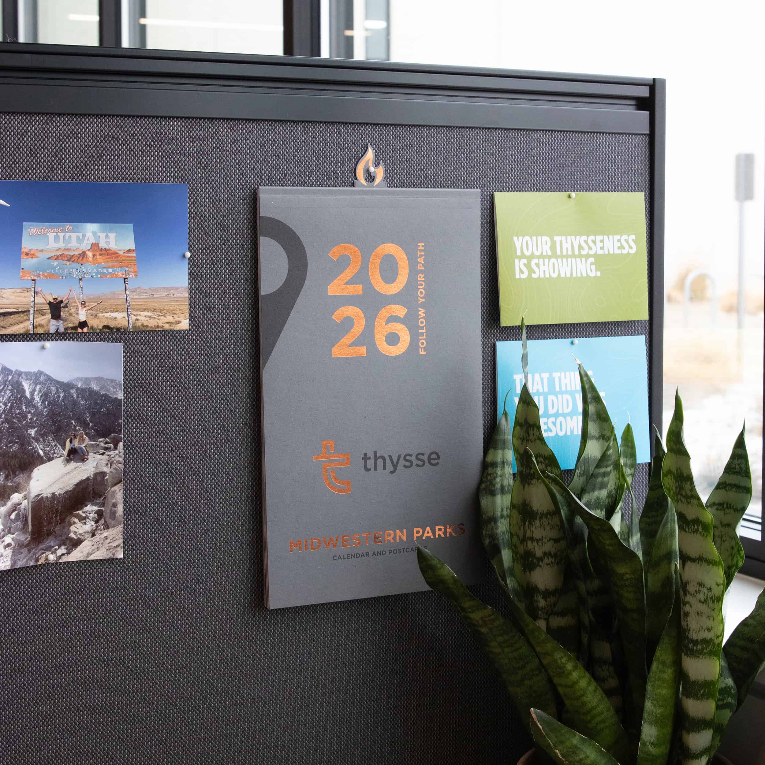 A Thysse employee workspace with the 2026 Midwestern Parks calendar displayed on a cubicle wall alongside postcards and brand cards. A potted snake plant, Thysse business card, branded “glow” box, and a “Made in the Midwest” mug sit on the desk near a window.