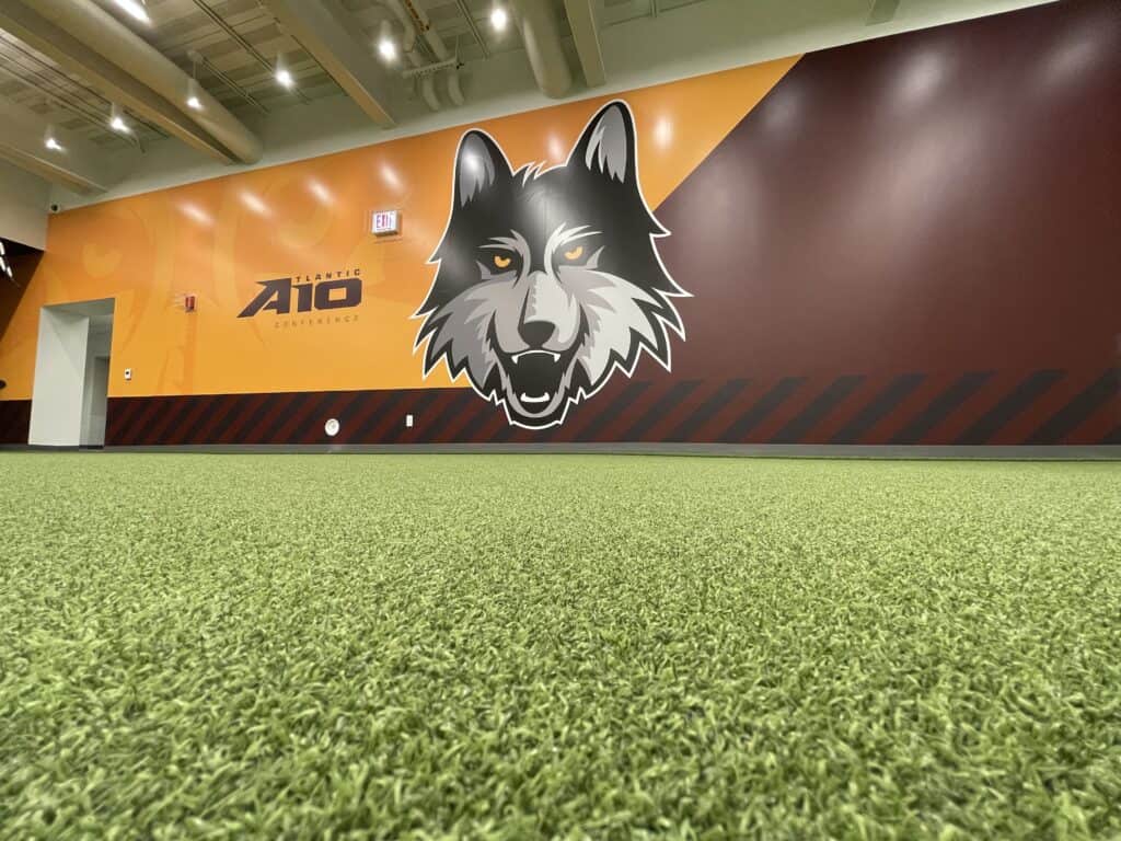 A full-wall wolf graphic and Loyola’s conference branding anchor the strength and conditioning room, set against turf flooring and bold color blocking.