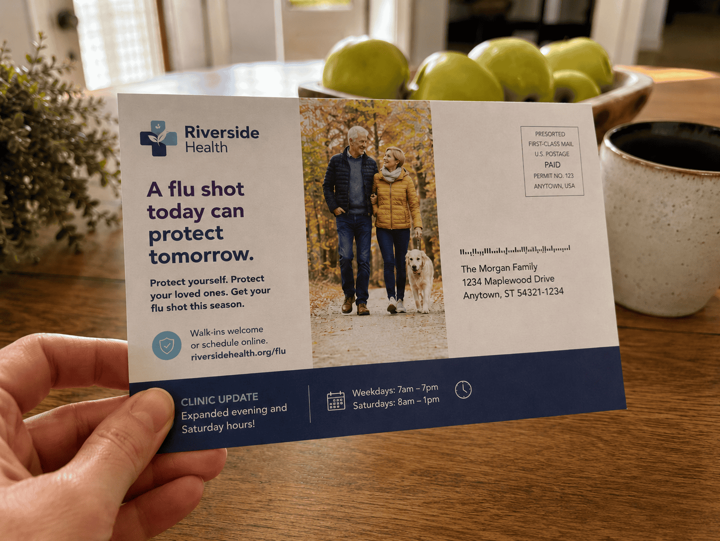 A First-Class Mail postcard from a healthcare organization promotes a flu shot reminder and clinic update while being held in a home kitchen setting.