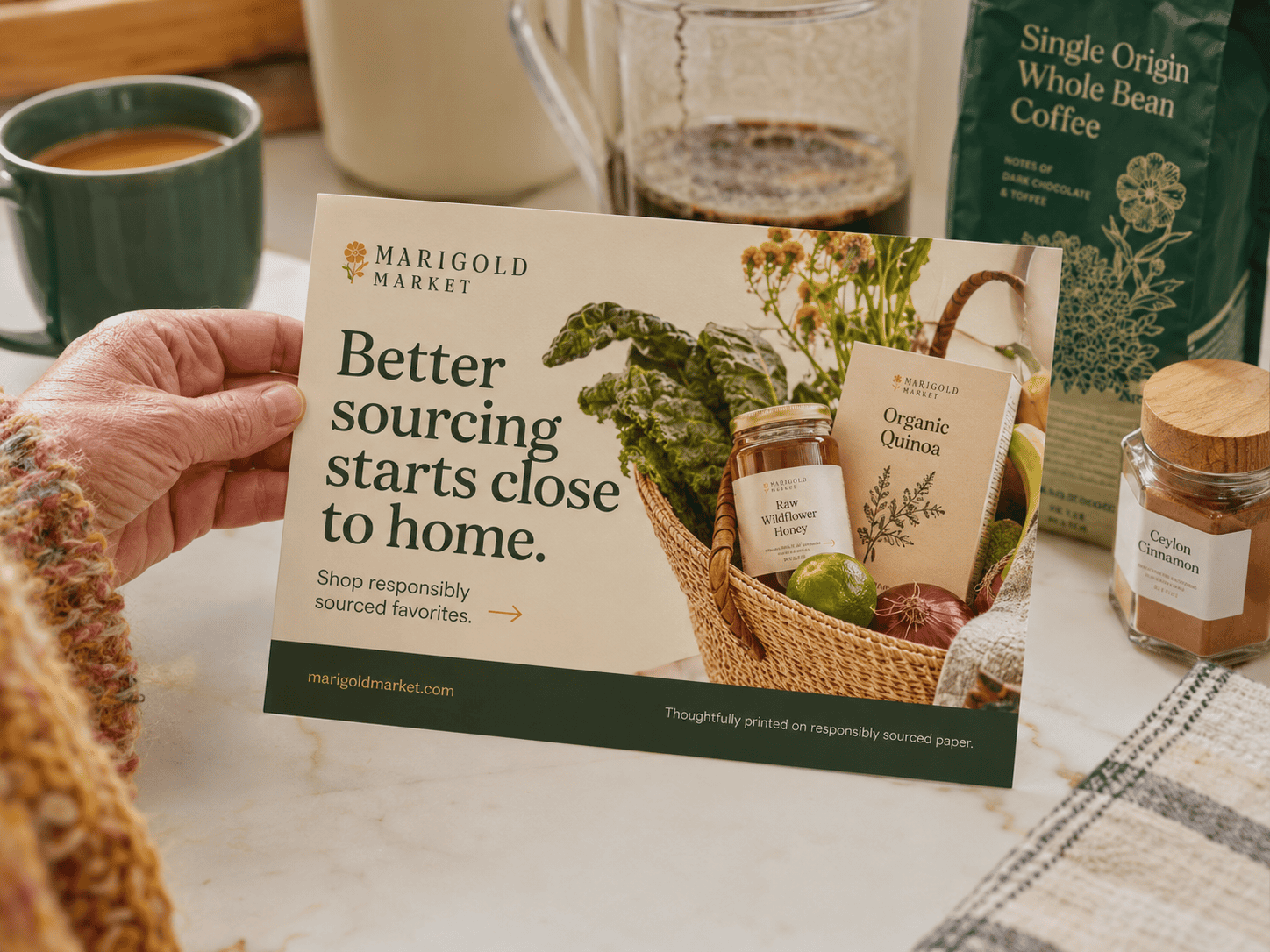 A person holding a Marigold Market postcard in a bright kitchen beside coffee, pantry goods and responsibly sourced products. The postcard promotes responsibly sourced favorites and includes a small footer note that it was thoughtfully printed on responsibly sourced paper.