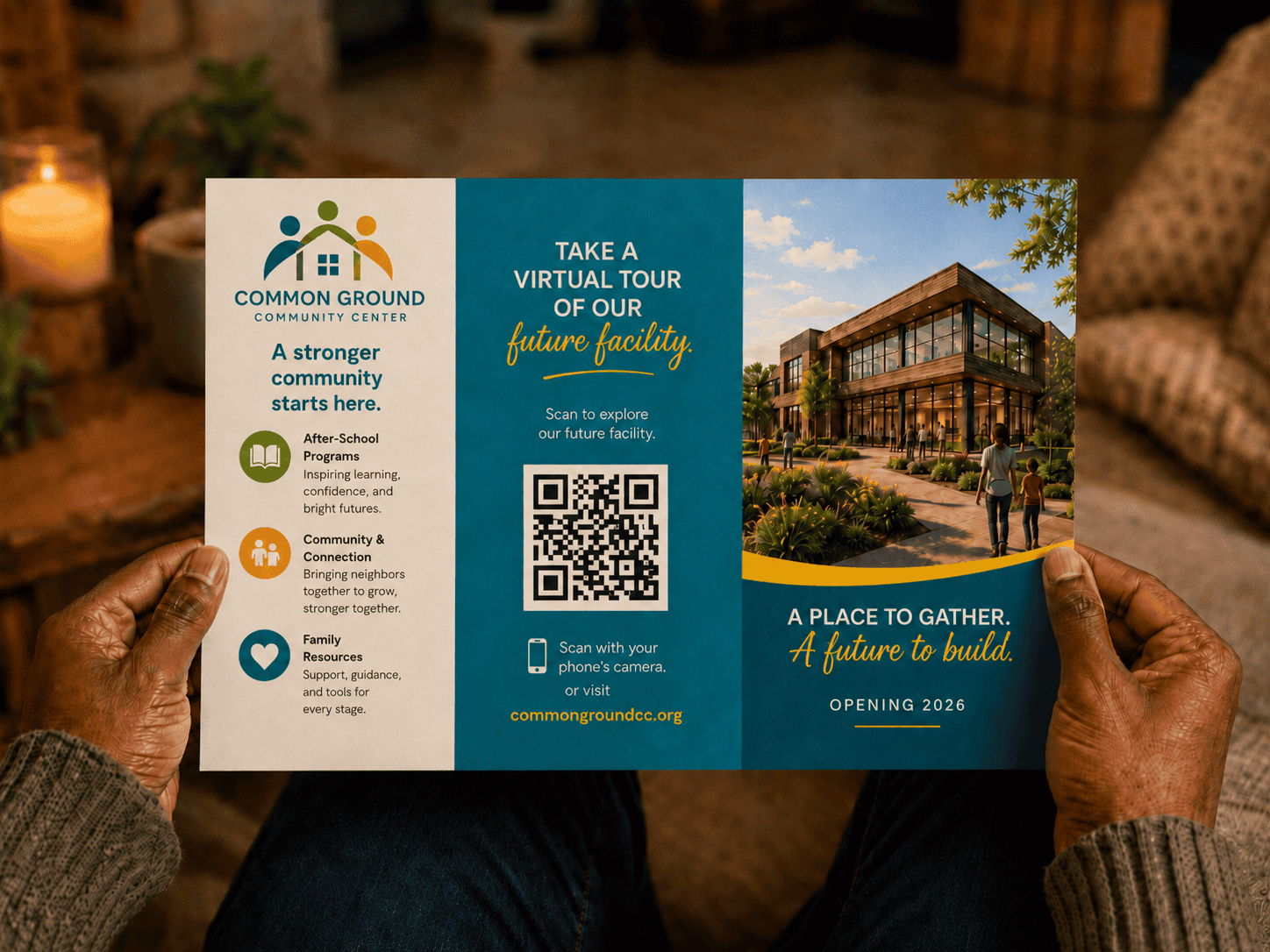 A person holding an opened double gate self-mailer for Common Ground Community Center in a cozy home setting. The mailer features a QR code inviting recipients to take a virtual tour of the future facility, with community program details, a building rendering and opening 2026 message.