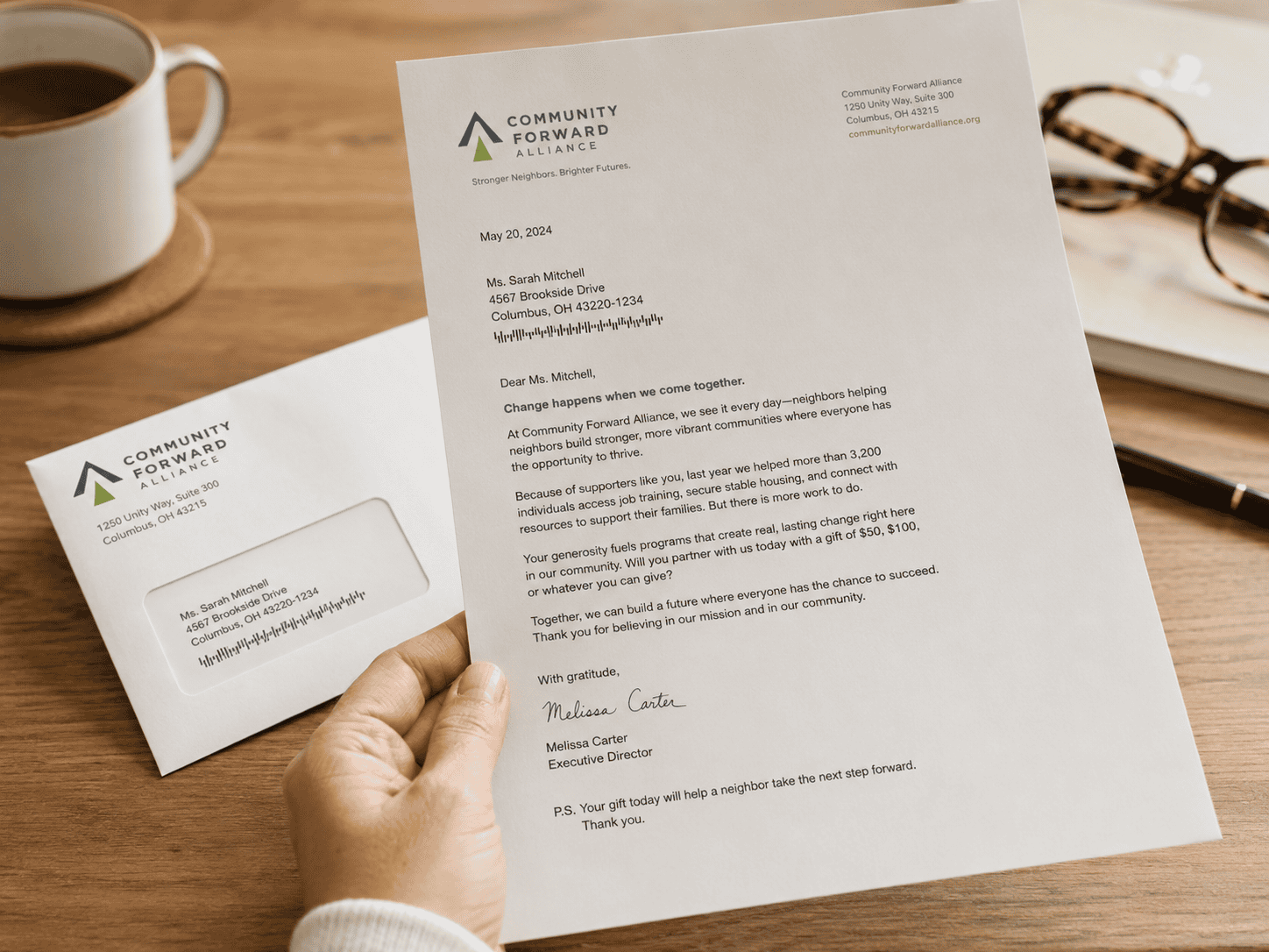 A personalized nonprofit fundraising letter signed by an executive director is held above a desk beside a matching envelope, showing a donor acquisition or membership appeal.